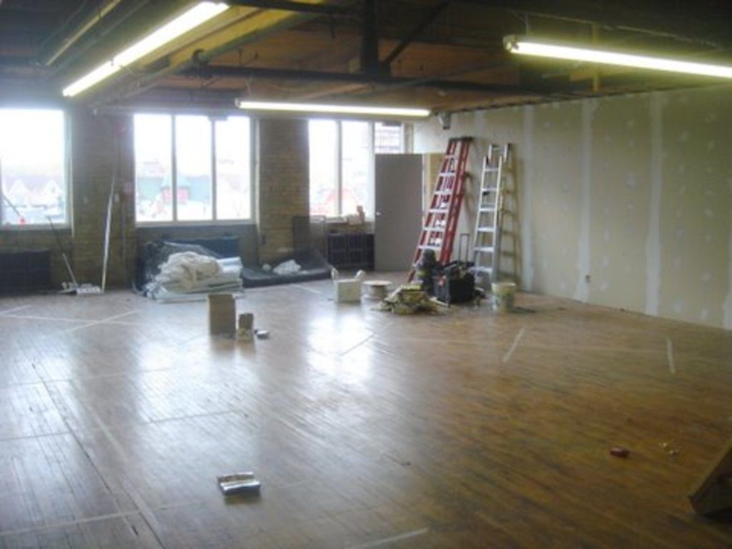 A large open room mid renovation with worn hardwood floors, ladders and drywall panels
